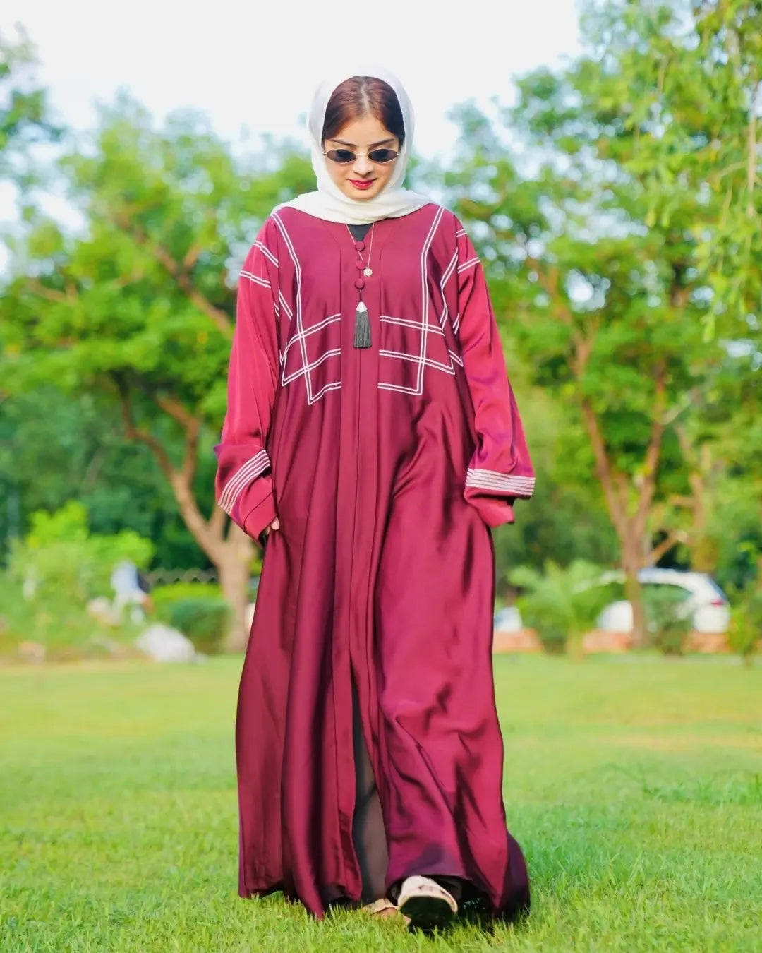 Woman in a burgundy abaya with white details standing in a park.