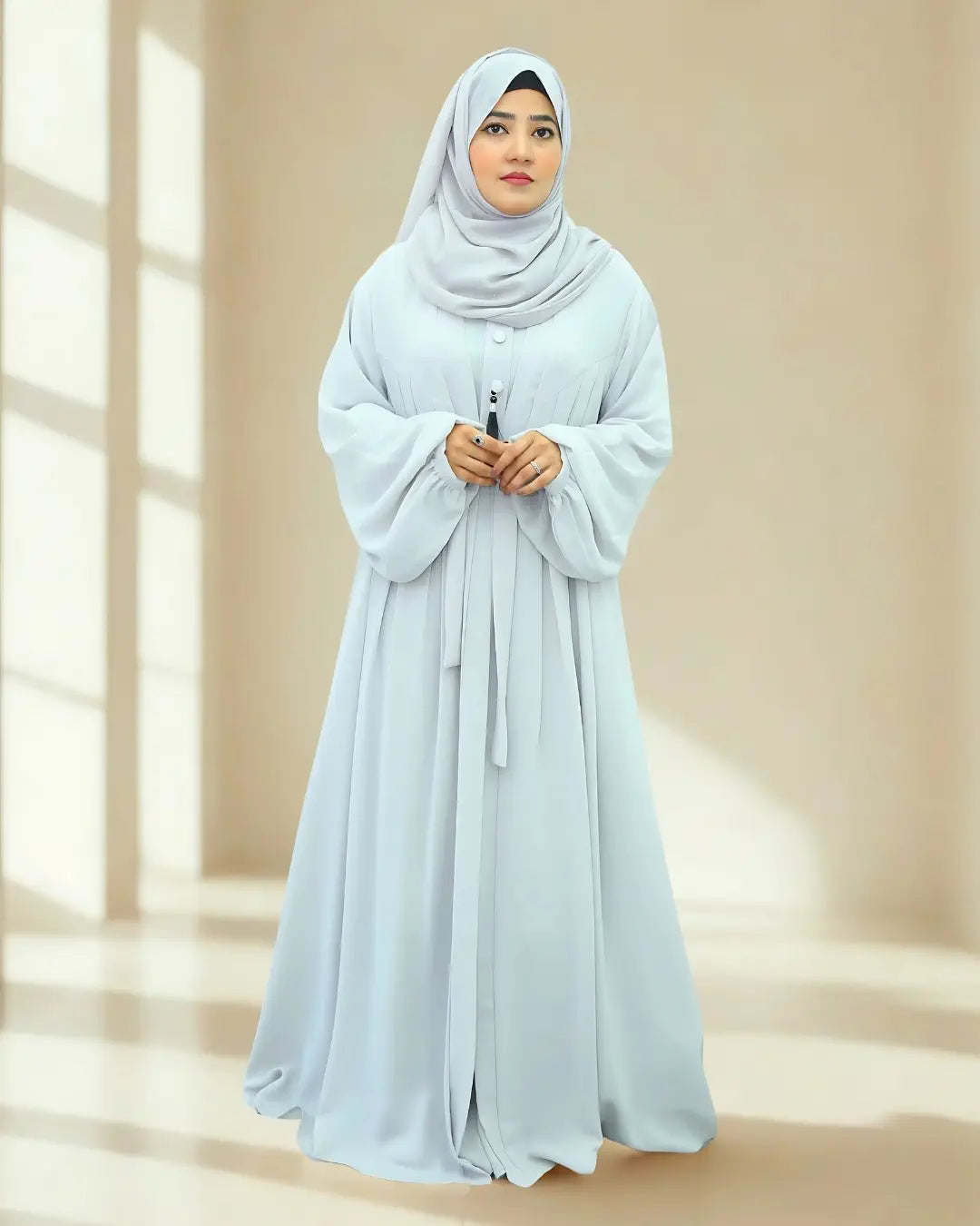 Woman wearing a light blue hijab and abaya in a bright room with natural light.