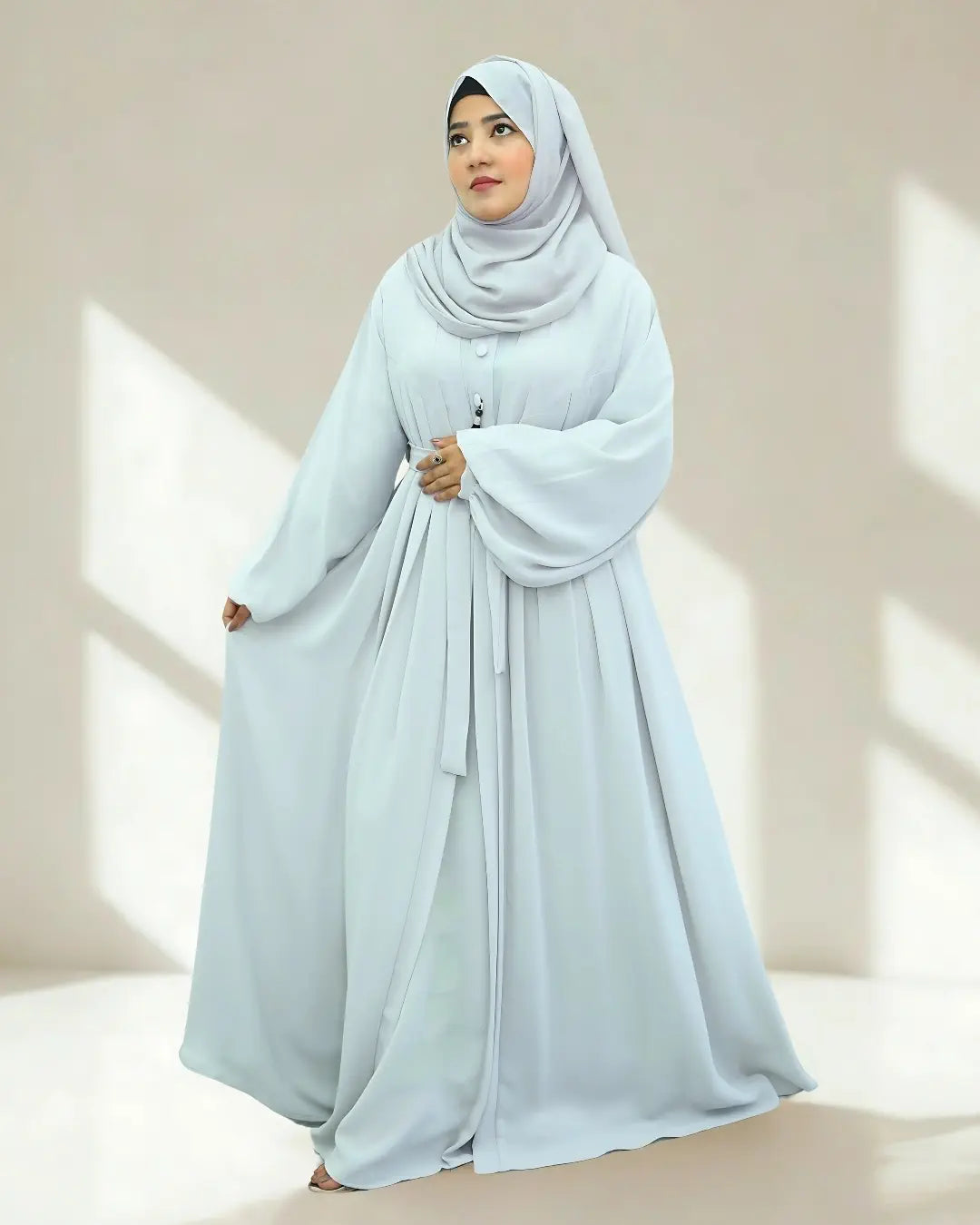 Woman wearing a light blue hijab and abaya in a bright room with natural light.