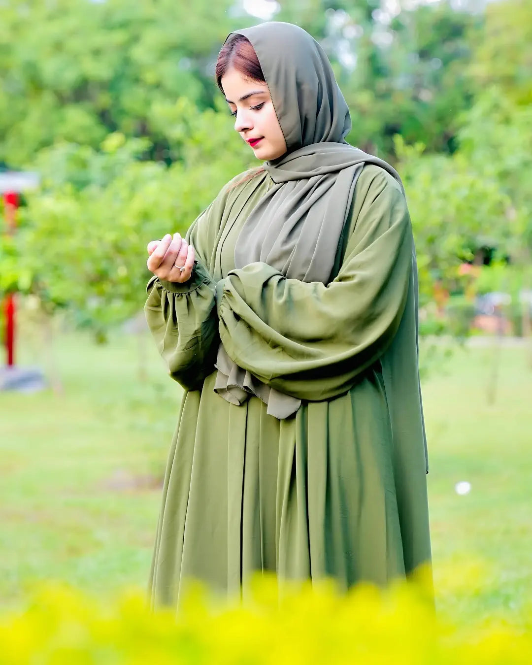 Woman in a green hijab and abaya standing in a grassy area with trees in the background