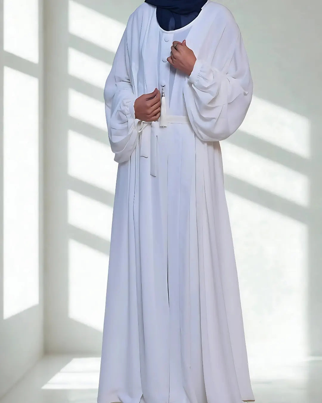 Person wearing a long white abaya with a blue headscarf in a bright room.