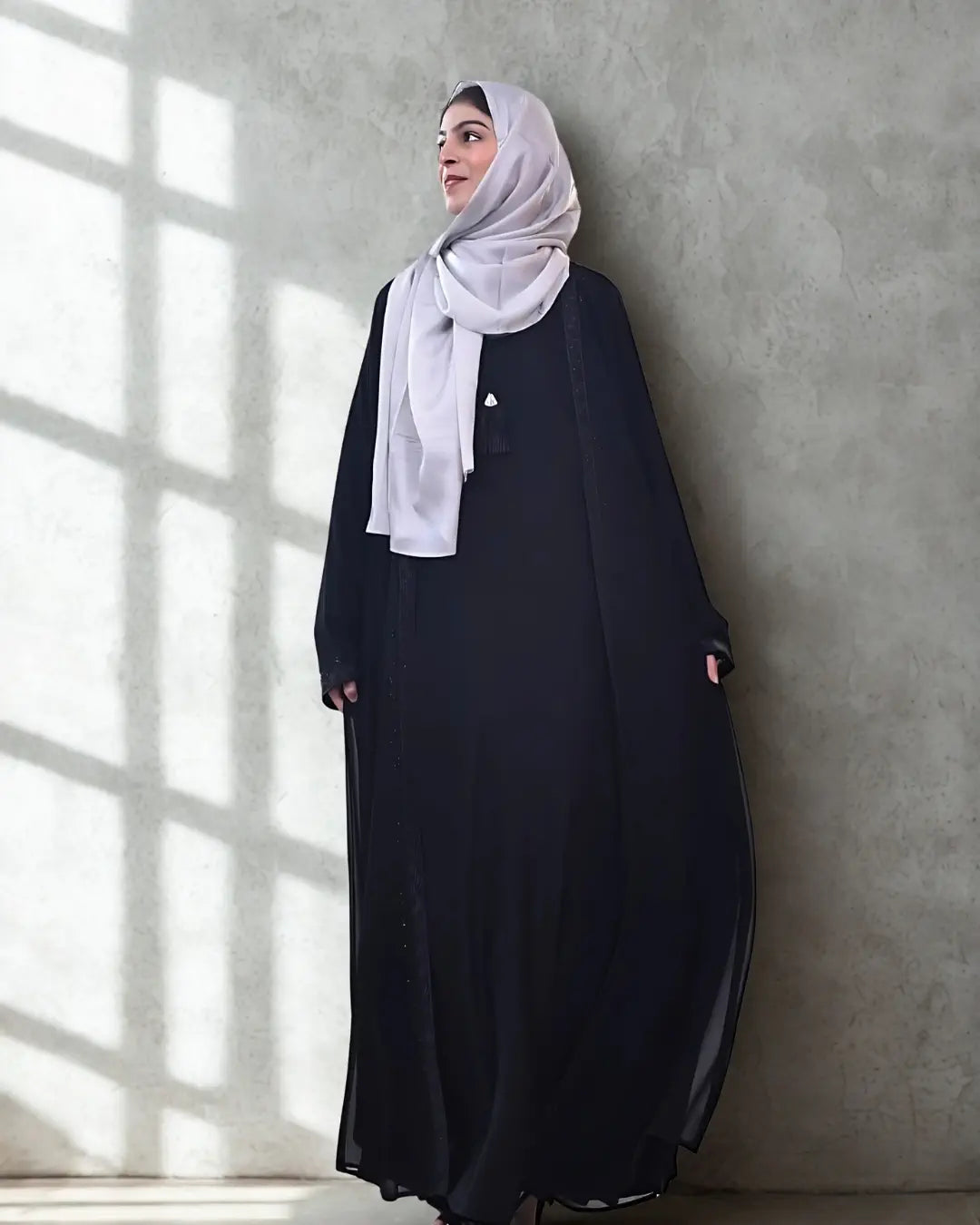 Woman wearing a black abaya and white hijab against a gray wall with sunlight casting shadows.