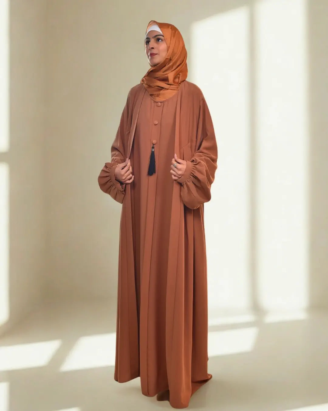 Woman wearing a long brown abaya and hijab in a bright room.