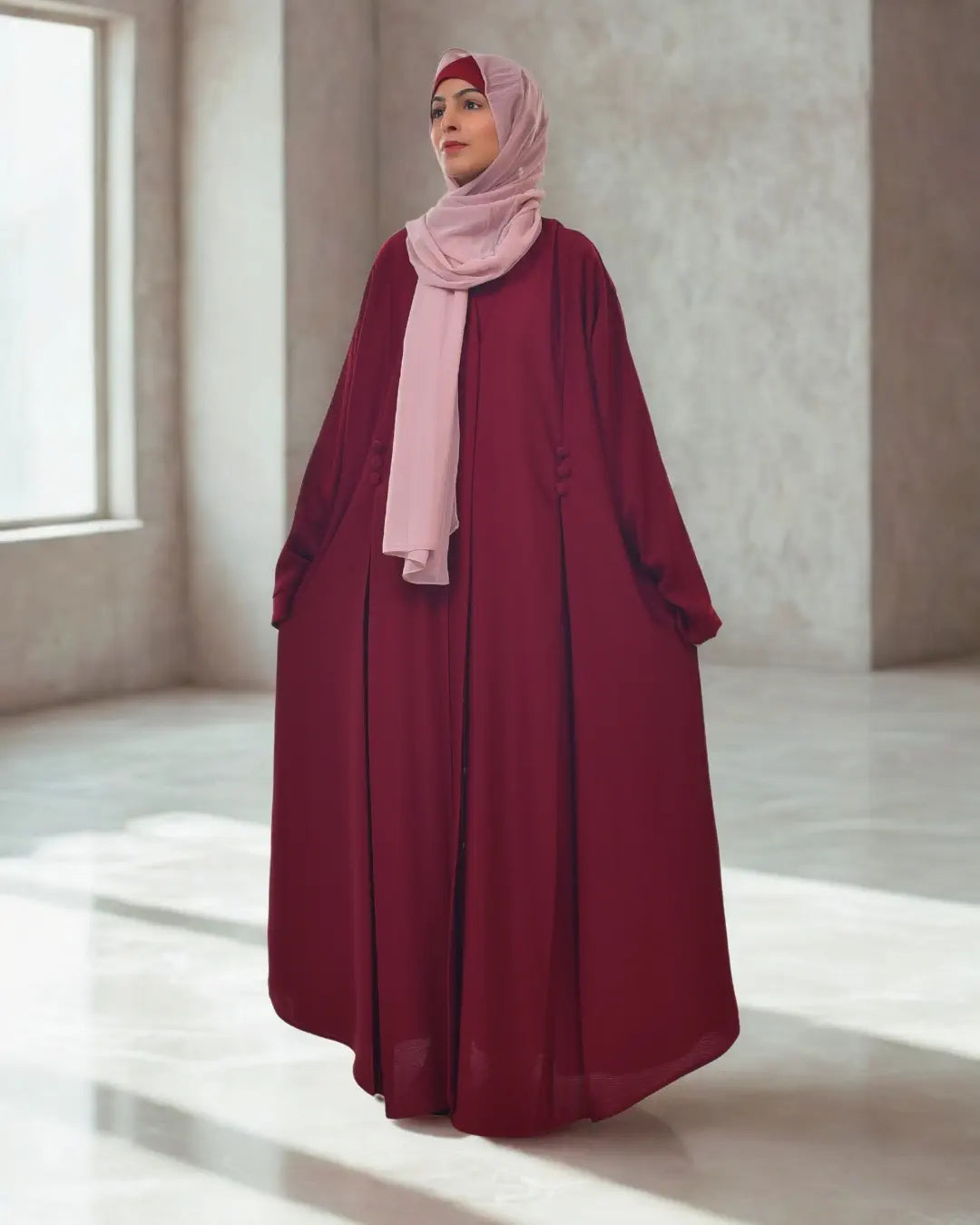 Woman wearing a burgundy abaya and pink hijab in a minimalistic room.