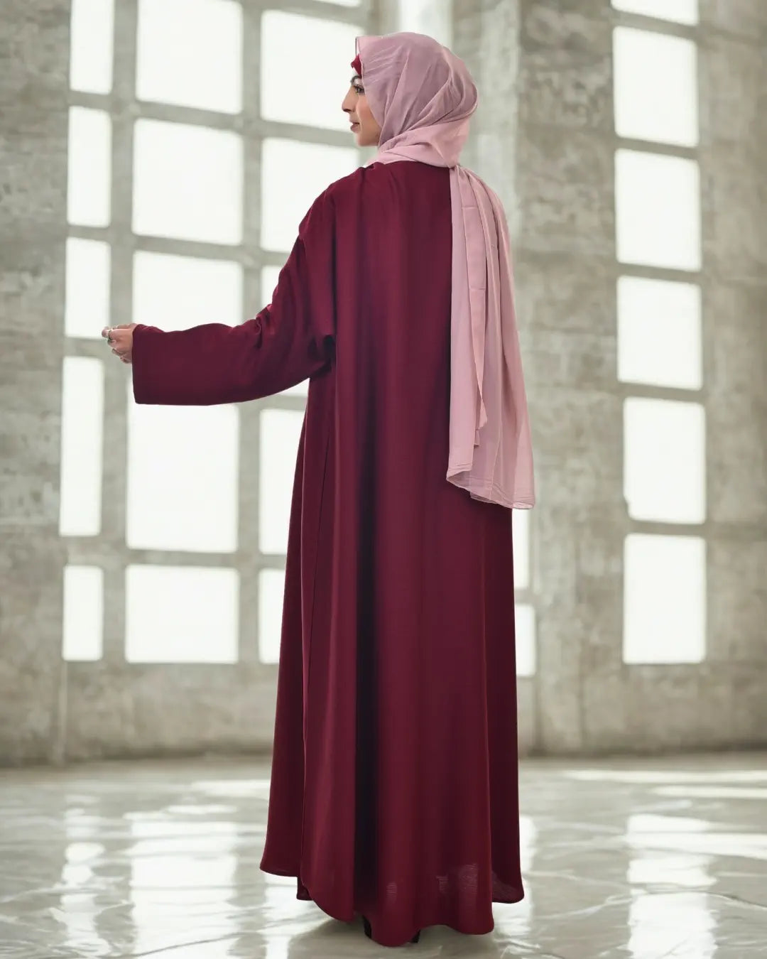 Woman wearing a burgundy abaya with a pink hijab in a bright room.