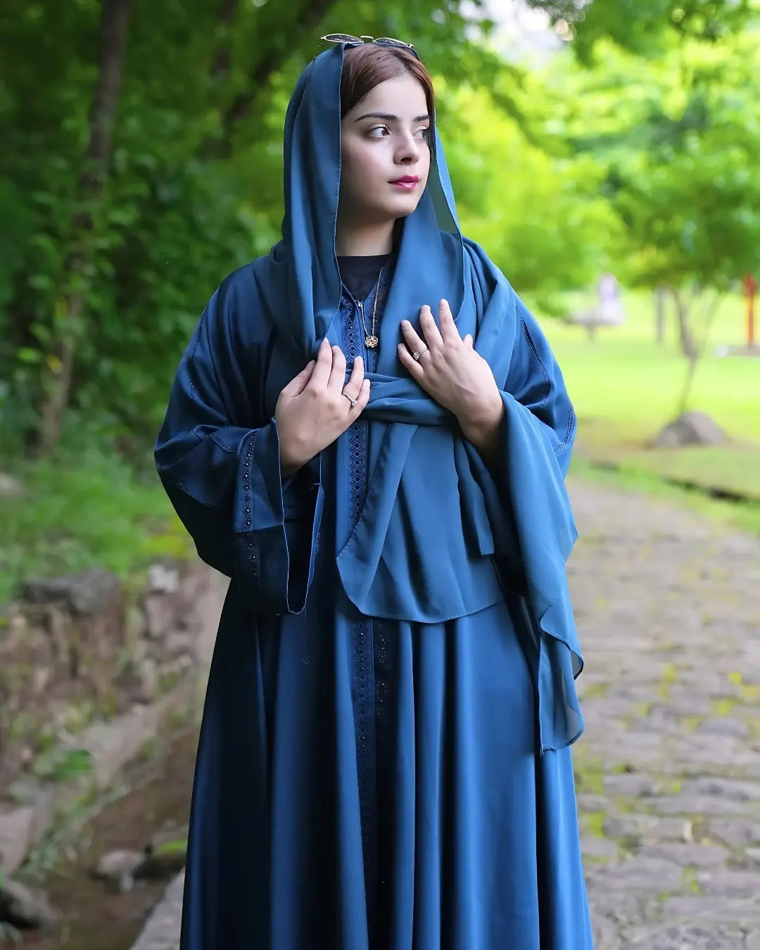 Woman in a blue abaya with same color hijab standing outdoors with greenery in the background