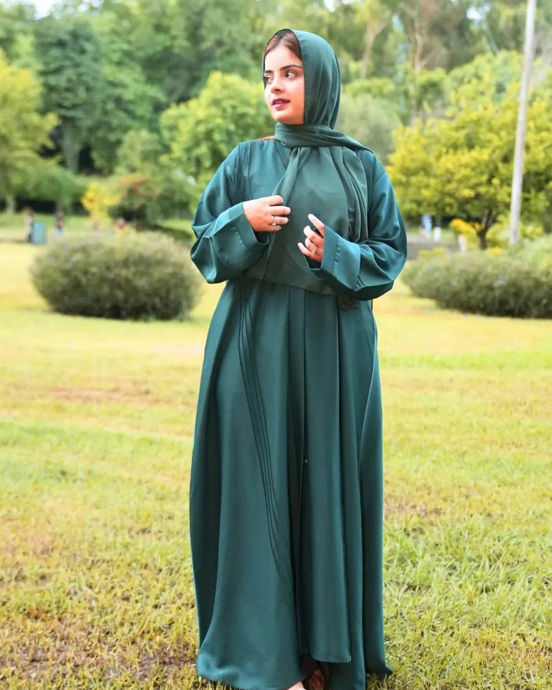 Woman in a green abaya standing in a park with trees and grass in the background