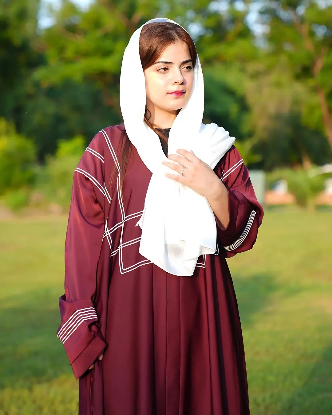 Woman in a burgundy abaya with a white headscarf standing on grass