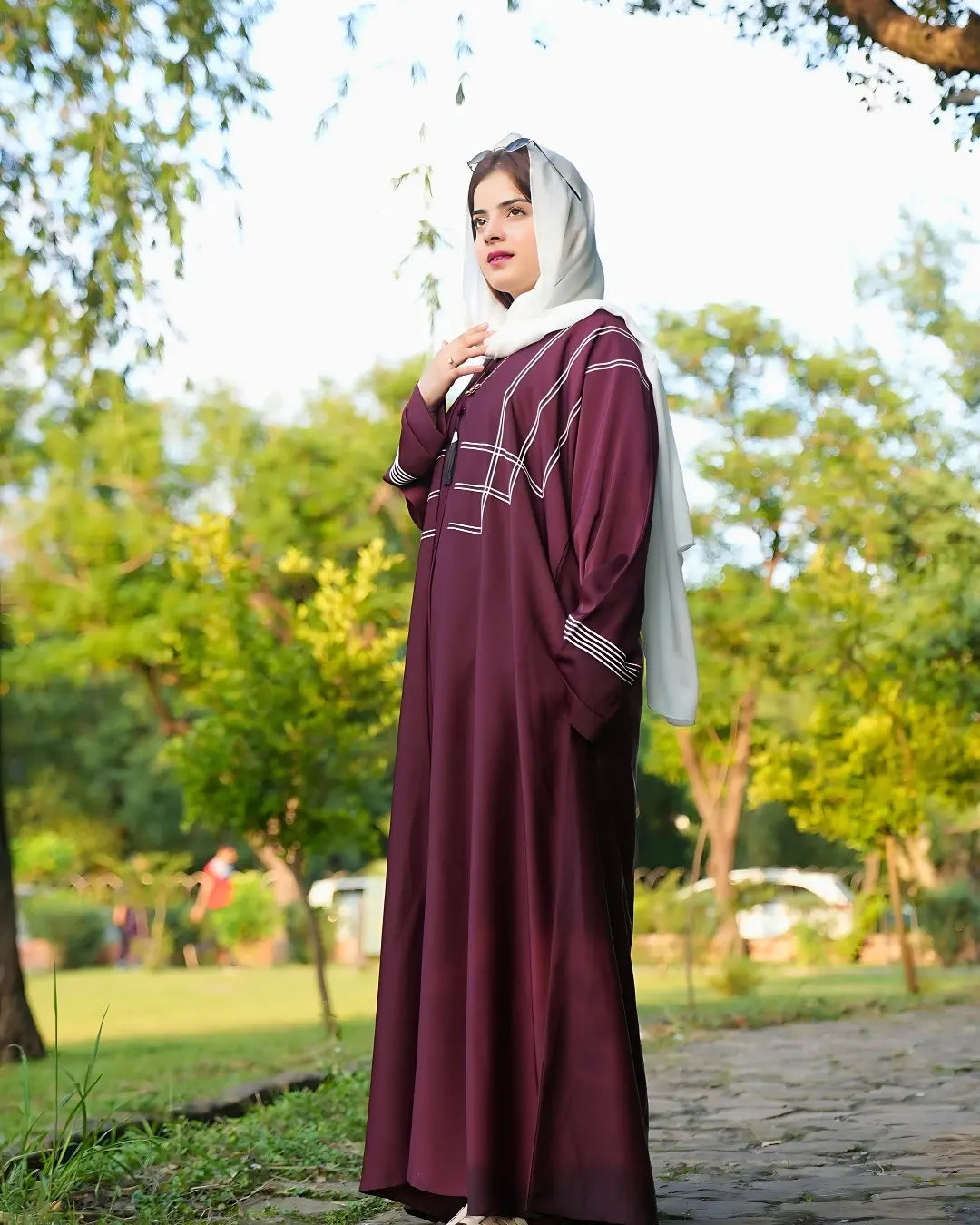 Woman in a burgundy abaya with a white hijab standing outdoors in a park.