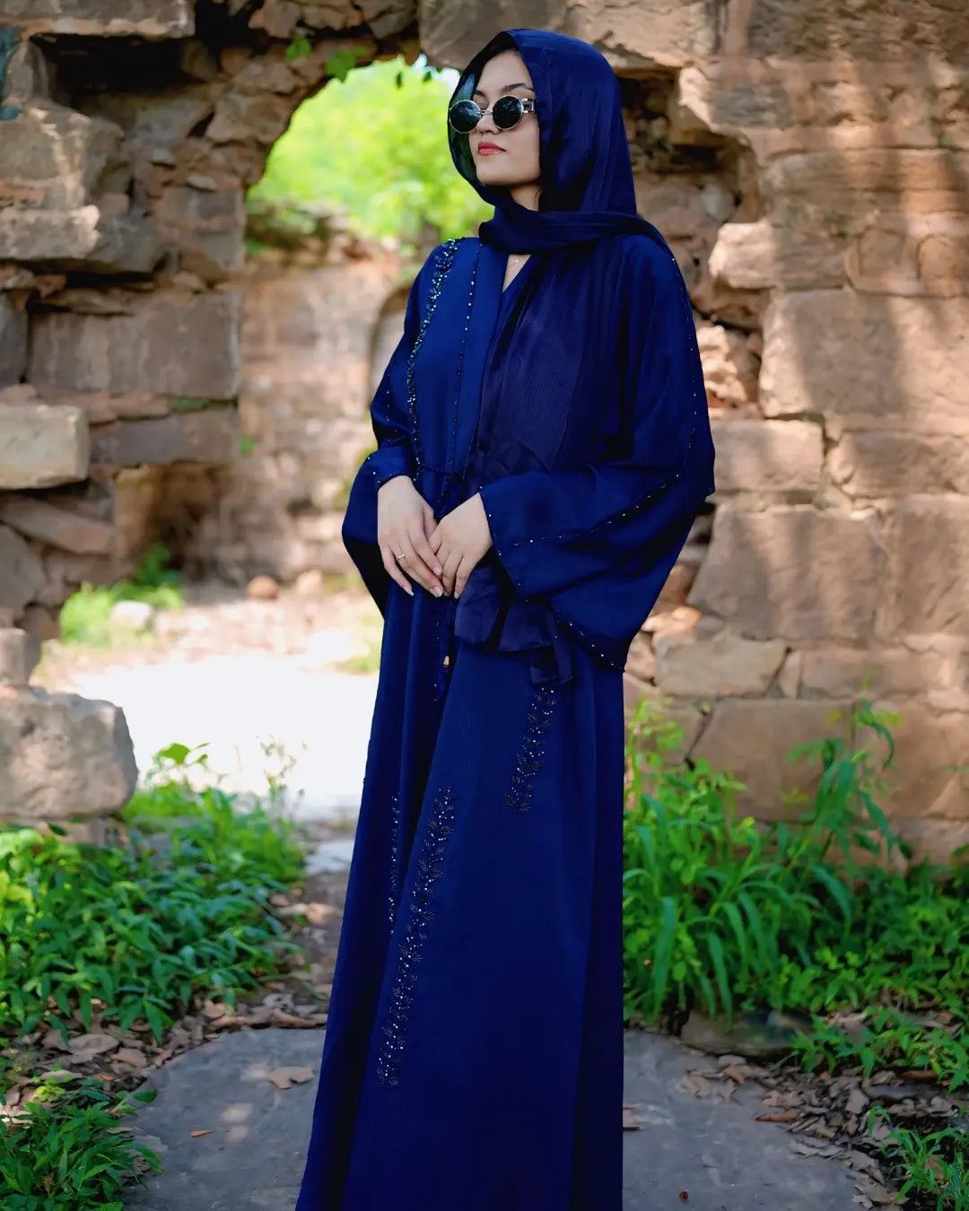 Woman in a blue abaya standing in front of stone ruins with greenery.