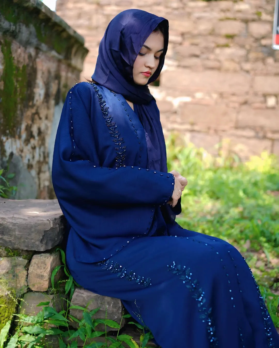 Woman in a blue abaya with intricate designs sitting outdoors.