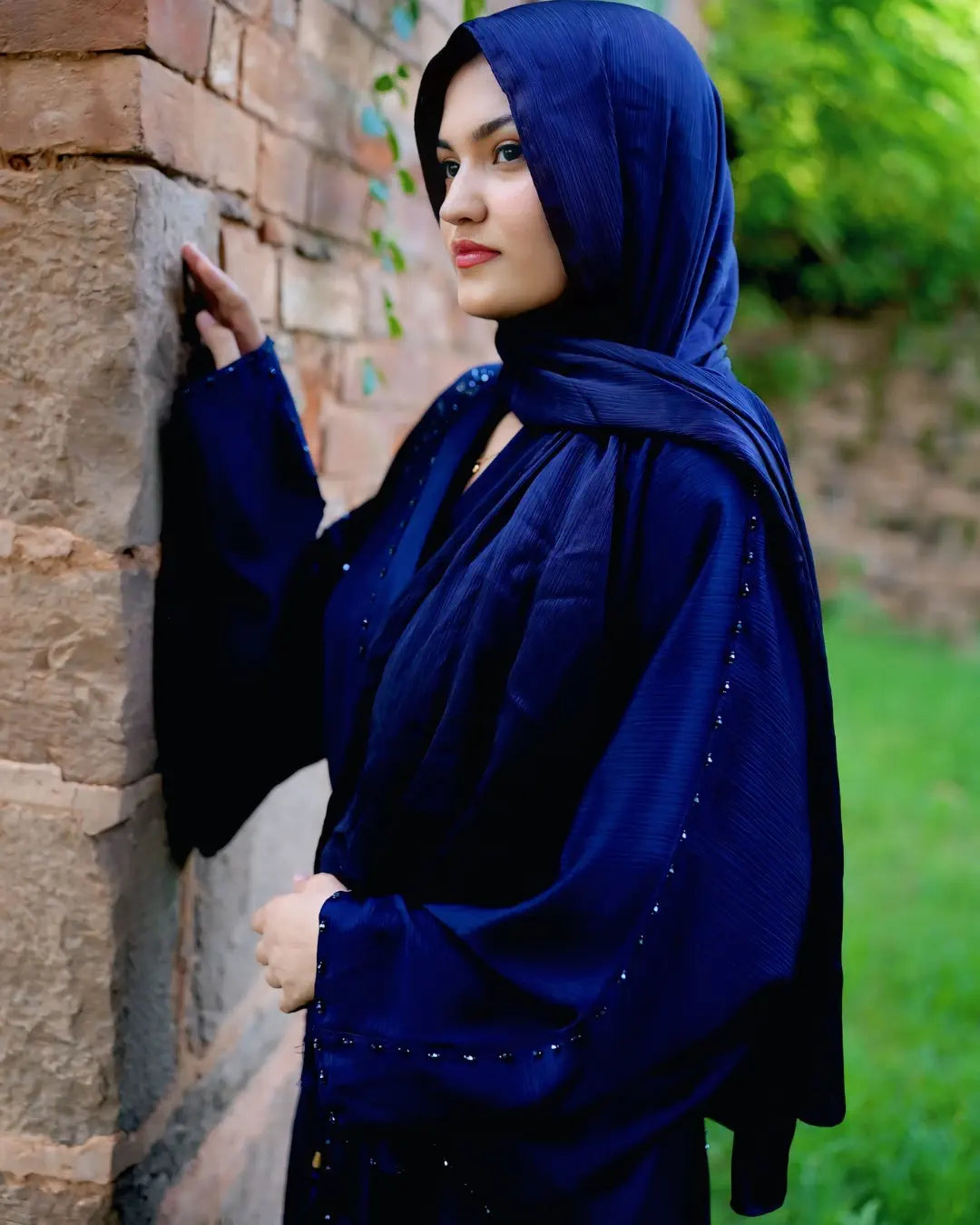 Woman wearing a blue hijab and abaya against a stone wall with greenery.