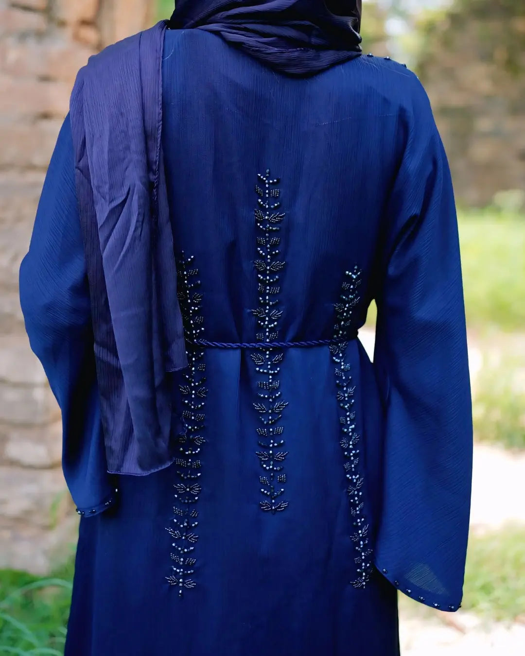 Navy blue abaya with intricate detailing on the back, worn by a person, against a blurred natural background.