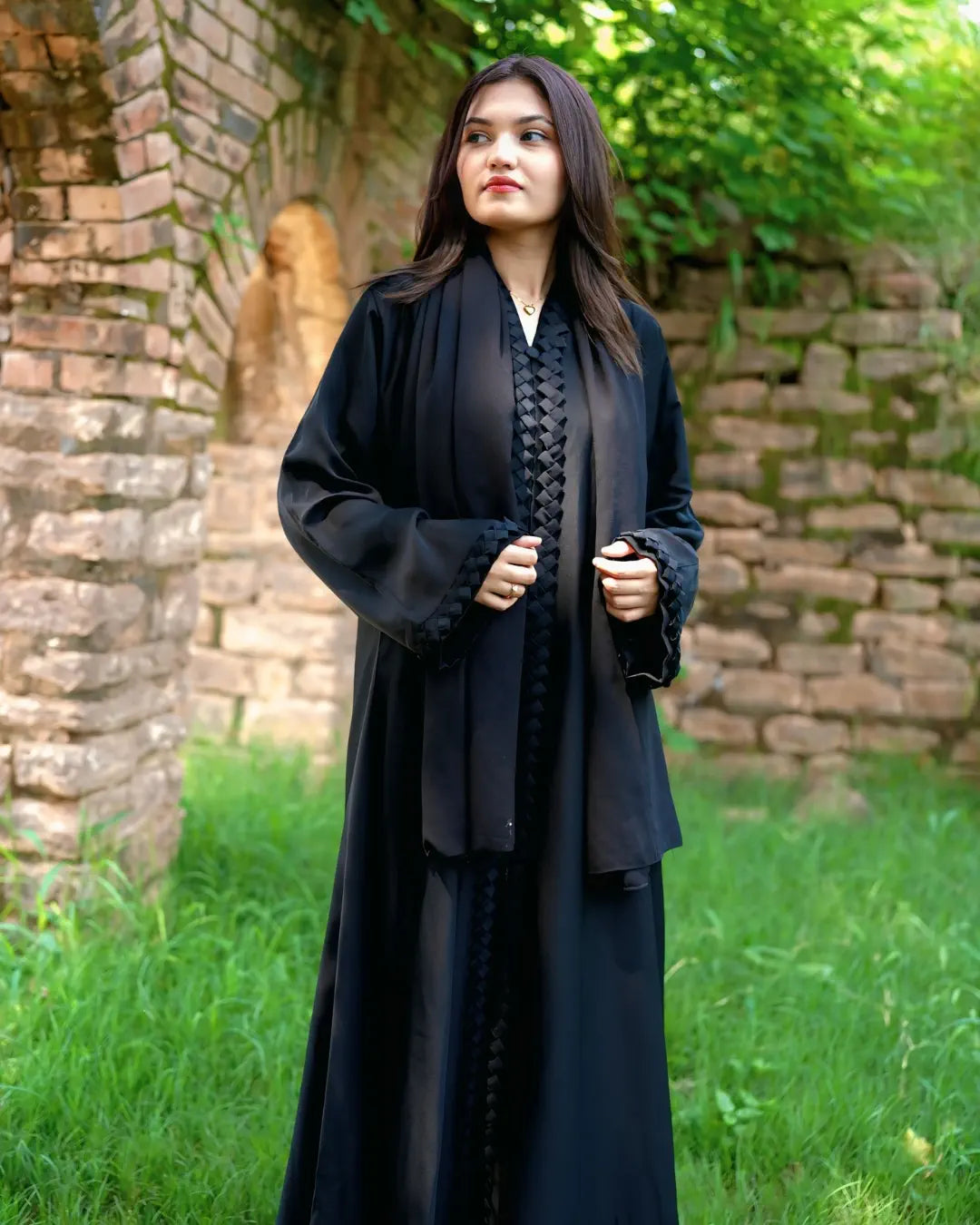 Woman in a black abaya standing outdoors with greenery and a stone wall in the background