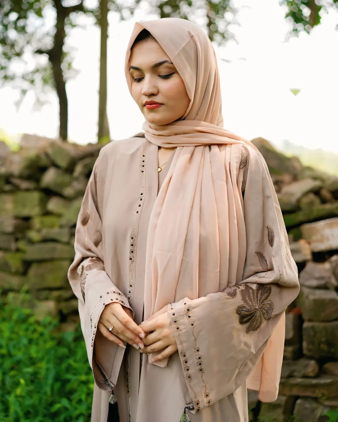 Woman wearing a beige hijab and abaya with a stone wall and greenery background