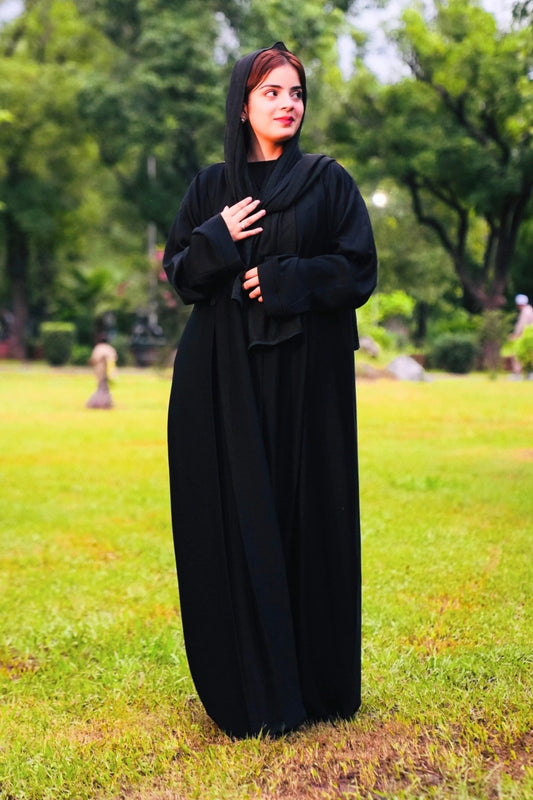 Woman in a black abaya standing in a park with green grass and trees.