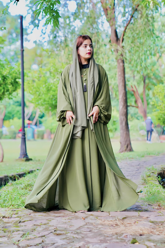 A girl in a green Abaya with scarf standing in a park with trees and people in the background