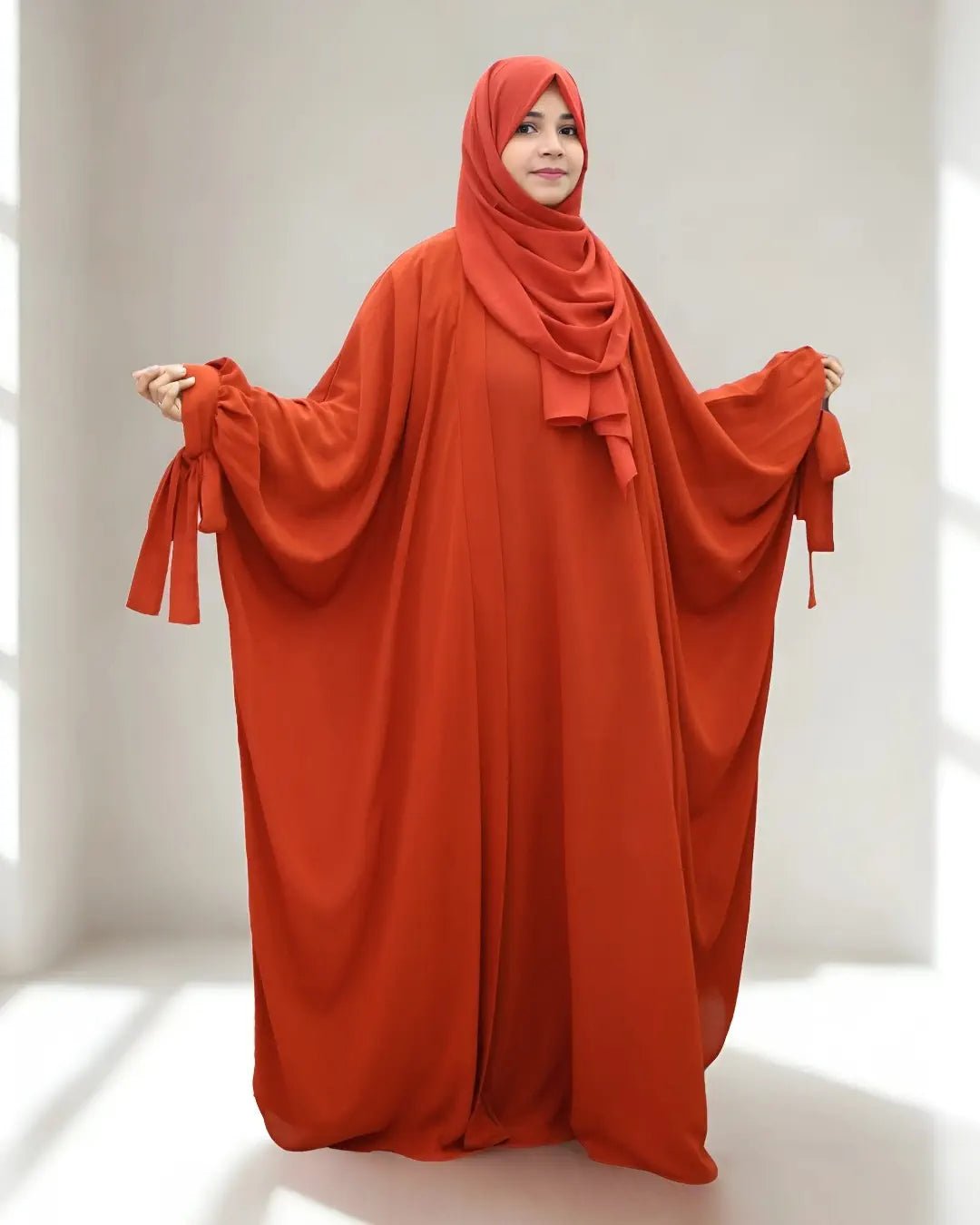 Person wearing a long, flowing orange abaya with a matching hijab in a bright room.