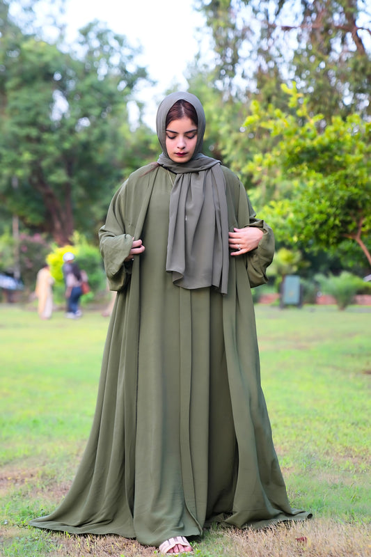 A girl in a green Abaya with scarf standing in a park with trees and people in the background