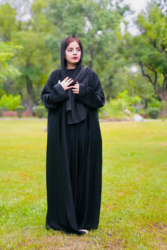 Person wearing a black hooded outfit standing in a grassy area with trees in the background