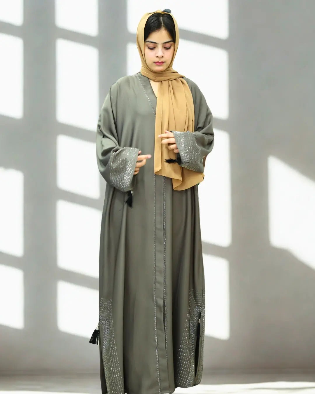 Woman wearing a long, light gray abaya with a matching hijab in a bright room with sunlight filtering through.