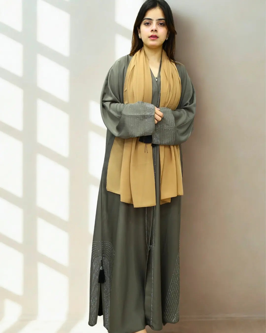 Woman wearing a long, light brown abaya with a gray cardigan against a neutral background
