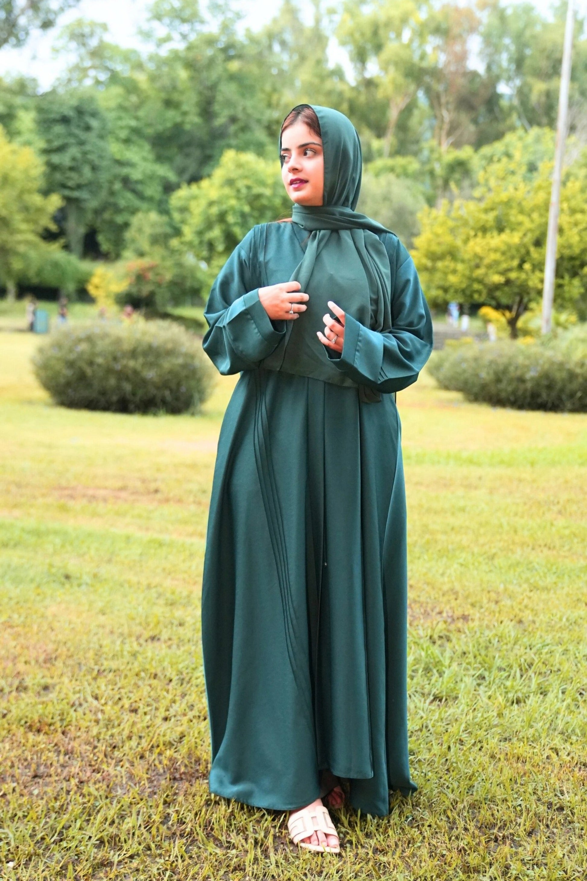 Woman in a green abaya standing in a park with trees and grass in the background