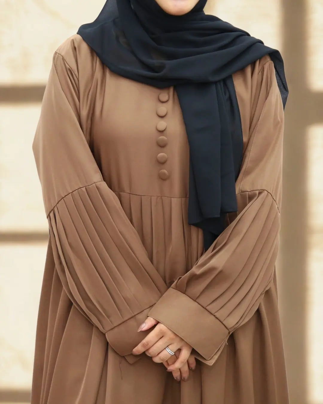 Person wearing a brown abaya with pleated sleeves and a black hijab on a neutral background