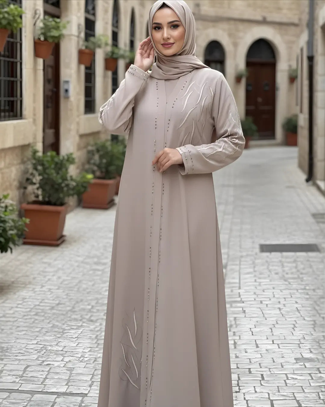 Woman in a long beige Abaya standing on a stone-paved street with buildings and plants in the background.