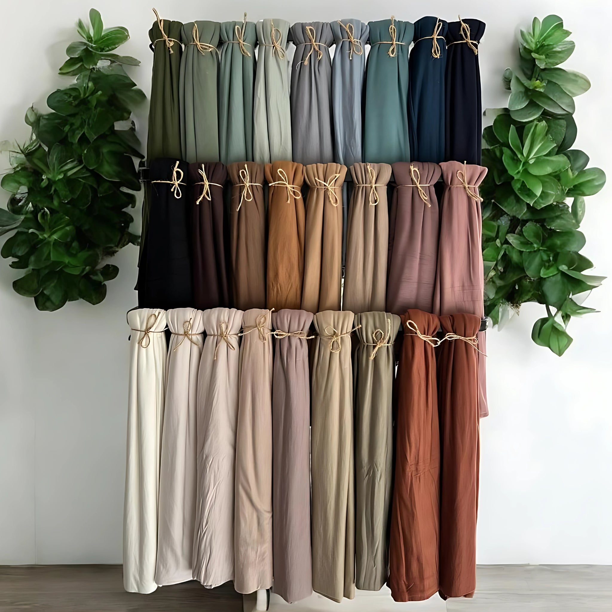 Collection of hijabs in various colors displayed against a white wall with green plants on either side.