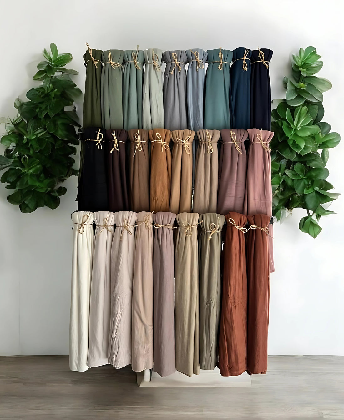 Collection of hijabs in various colors displayed against a white wall with green plants on either side.