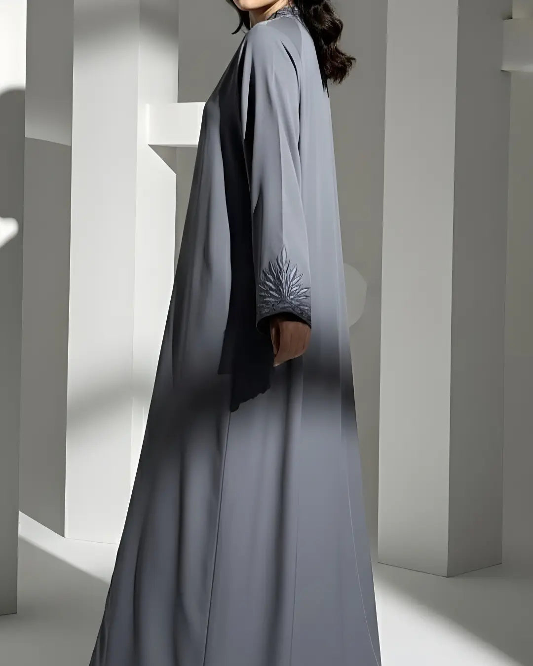 Person wearing a long, light gray ABAYA with a subtle pattern in a minimalistic setting.