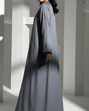 Person wearing a long, light gray ABAYA with a subtle pattern in a minimalistic setting.
